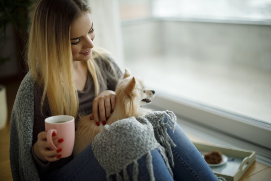 Kvinne som sitter i en vinduskarm med kaffekoppen i hånden og med en liten hund på fanget 